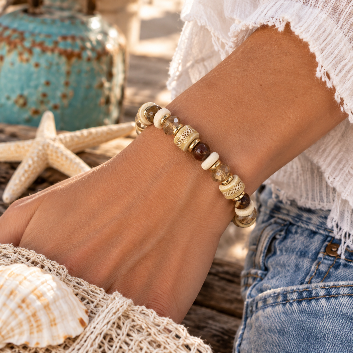 A close-up of a person's wrist wearing a beaded bracelet made of natural wood, cream and ivory beads, and textured ceramic beads. The person is wearing a white fringed top and blue jeans, with a seashell, starfish, and turquoise decorative pot in the background.