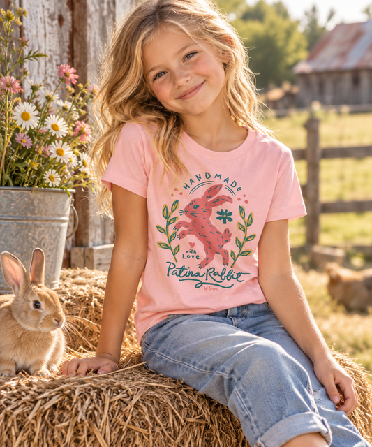 A young girl with blonde hair sits on a hay bale in a sunny outdoor farm setting, wearing a light pink graphic t-shirt with a rabbit design and blue jeans. A small brown rabbit is sitting next to her on the hay.